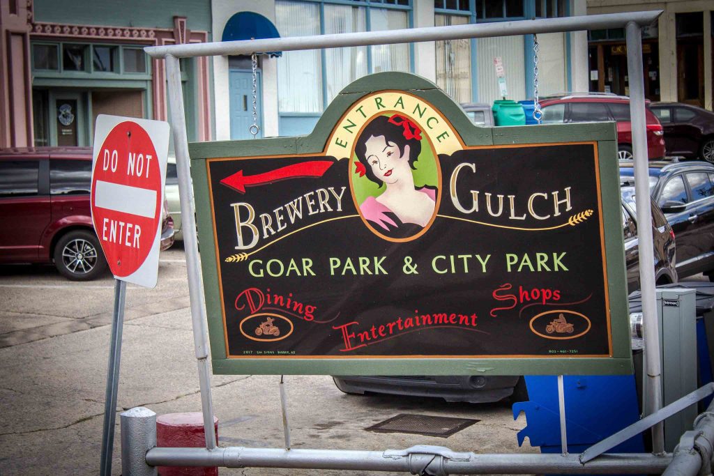 Outdoor sign with a painting of a woman, a red arrow, and the words “Entrance. Brewery Gulch. Goar Park & City Park. Dining Entertainment Shops,” next to a Do Not Enter sign.