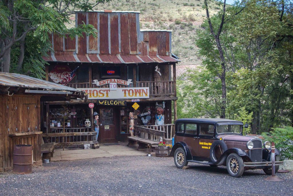 Brown old-timey car in front of a two-story wooden and metal building, with a sign above the entryway reading, “Entrance to Ghost Town. Welcome.”