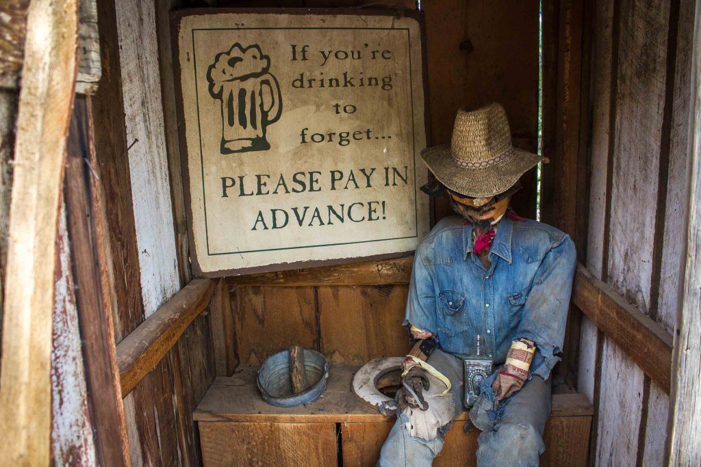 Interior of an outhouse with a mannequin dressed like a cowboy sitting next to the toilet seat, and a sign on the wall reading “If you’re drinking to forget…Please pay in advance!”