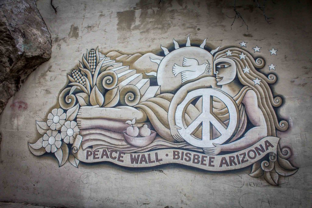 A mural of a reclining woman holding a white peace sign, above the words “Peace Wall Bisbee Arizona,” with a staircase, sun, and dove in the background.