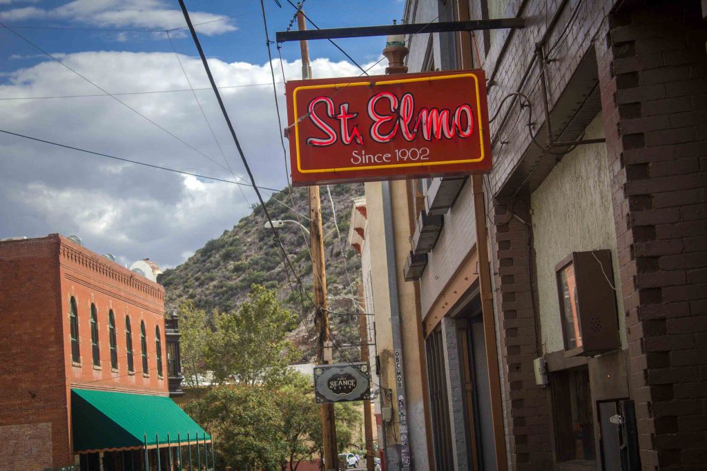 Red light-up sign reading “St. Elmo Since 1902,” mounted on the outside of a building, with a grassy hill in the background.