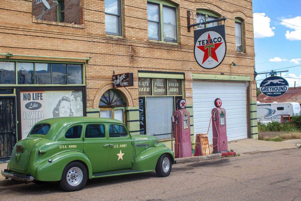 An old green car labeled U.S. Army, parked in front of two red Texaco gas pumps and a signed reading “Greyhound Lowell Arizona.”