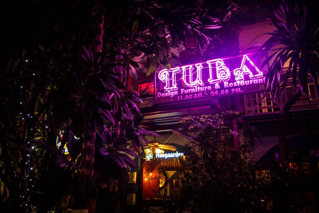Lit up purple neon sign that says Tuba Design, Furniture, and Restaurant amongst palm leaves.