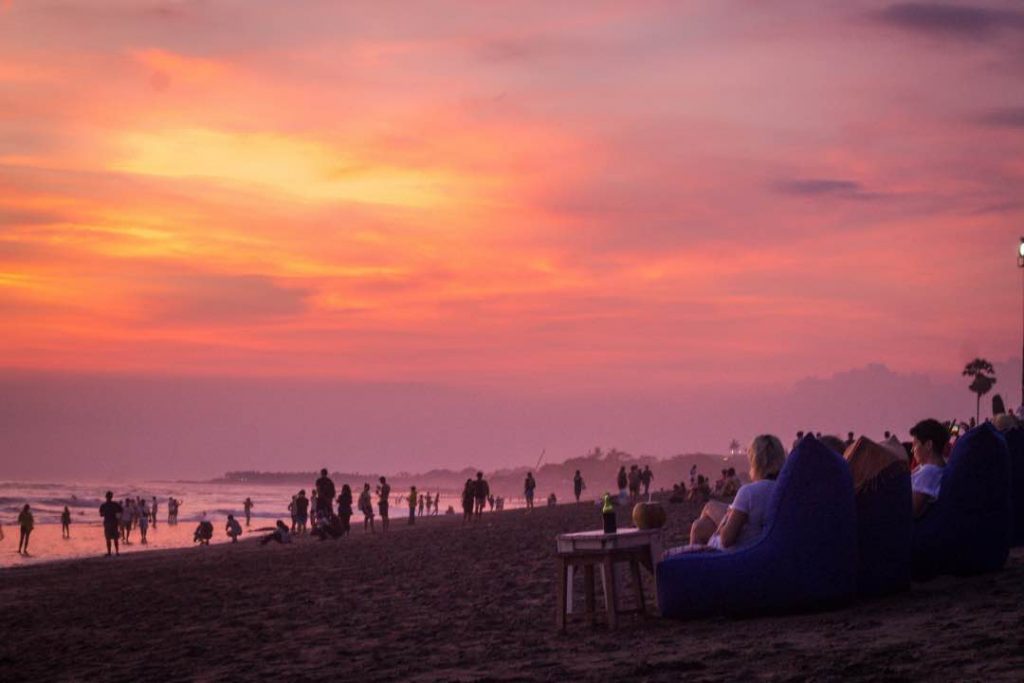 Crowd of people sitting in bean bag chairs on a beach watching a vibrant red sunset.