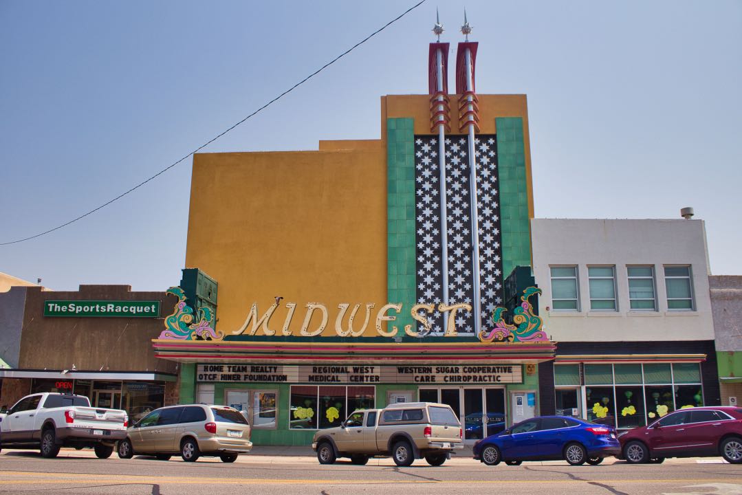 Art Deco theater with retro marquee stating "Midwest" with a line of cars angled parked in front of the building.