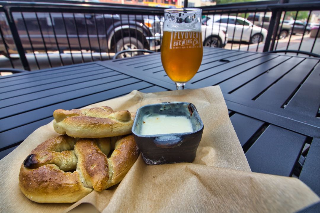A black metal table with a piece of butcher paper holding a soft pretzel and cheese deep and a goblet of copper-hued beer in the background.