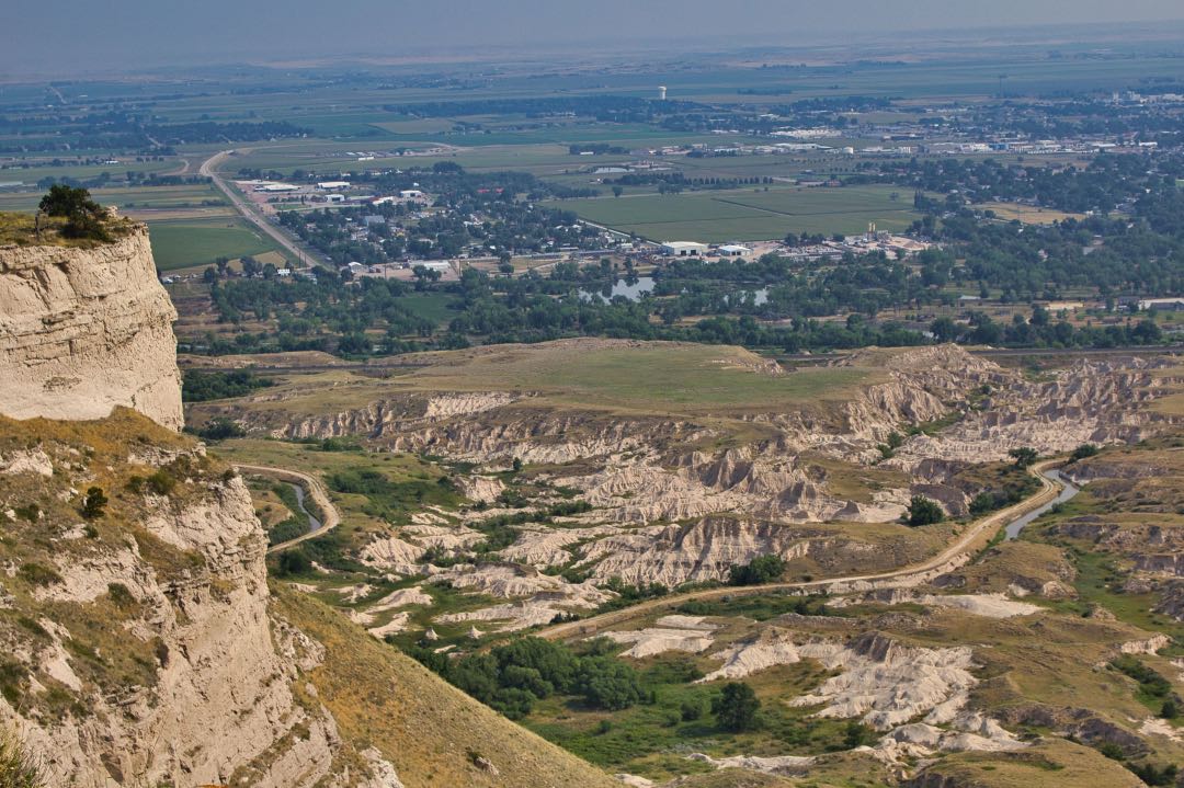 View from atop a tall rock formation with badlands and small cityscape in the background.