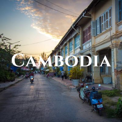"Cambodia" written across a photo of motorbikes in a narrow street, lined on one side with yellow buildings with colorful shutters.