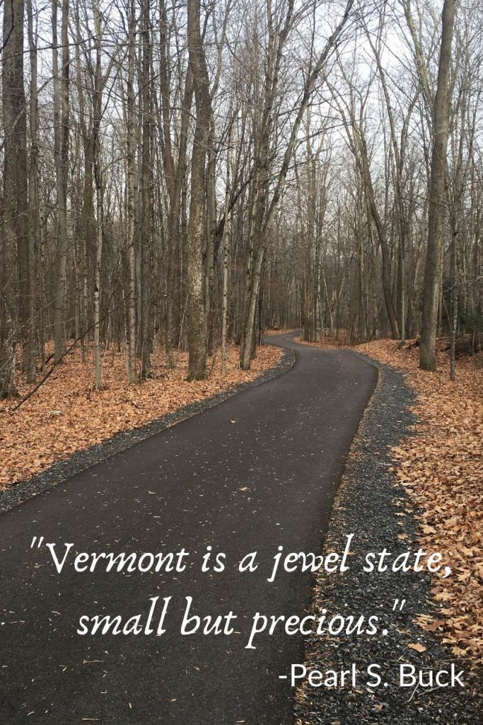 Asphalt path lined with bare trees and leaves on the ground, behind the quote, "Vermont is a jewel state, small but precious. -Pearl S. Buck."