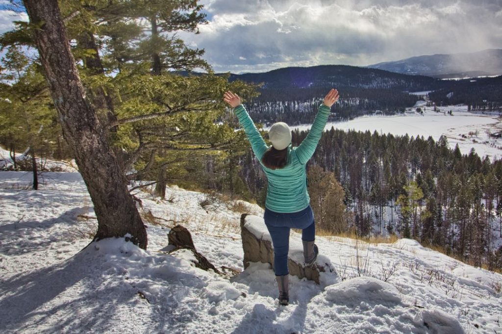 Woman in jeans, green puffy coat, and white hat standing in snow and looking out over forested hills.