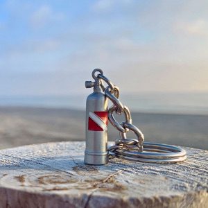Tiny silver scuba tank with red and white dive flag attached to a keyring, sitting on a wood at a beach.
