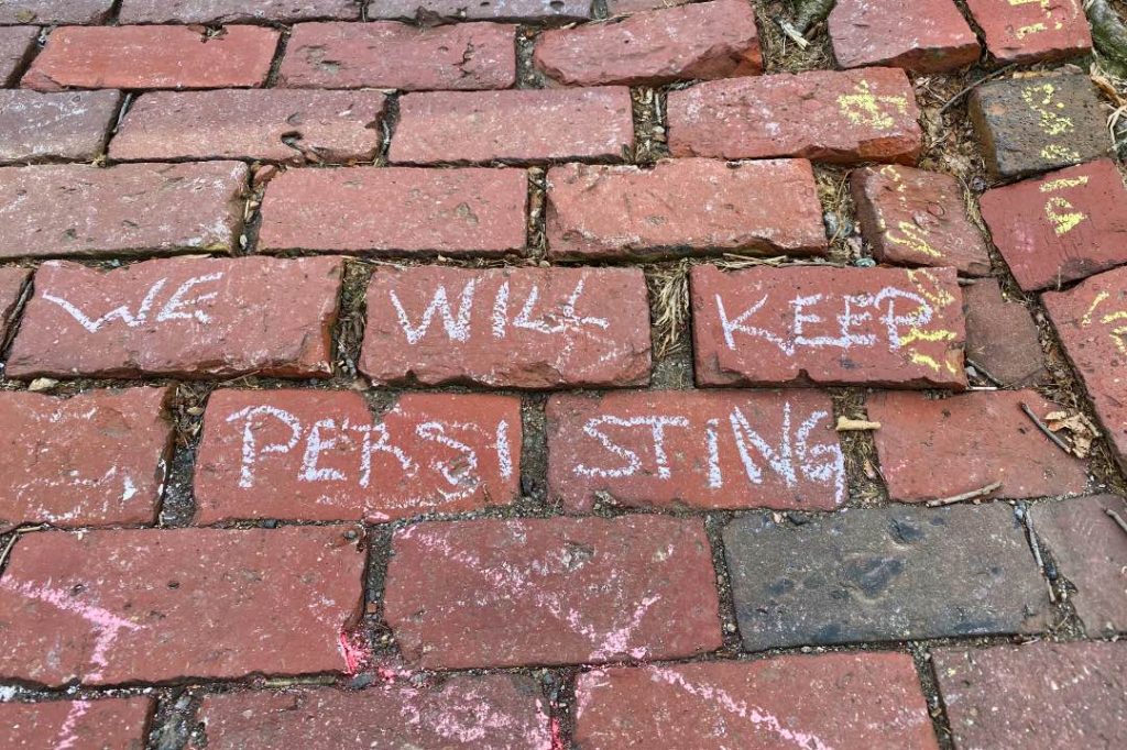 "We will keep persisting" written in white chalk on a brick sidewalk..