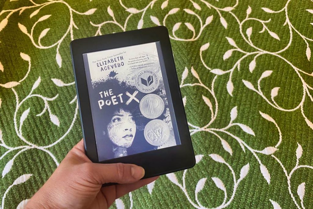 Hand holding a Kindle displaying the cover of "The Poet X" by Elizabeth Acevedo.