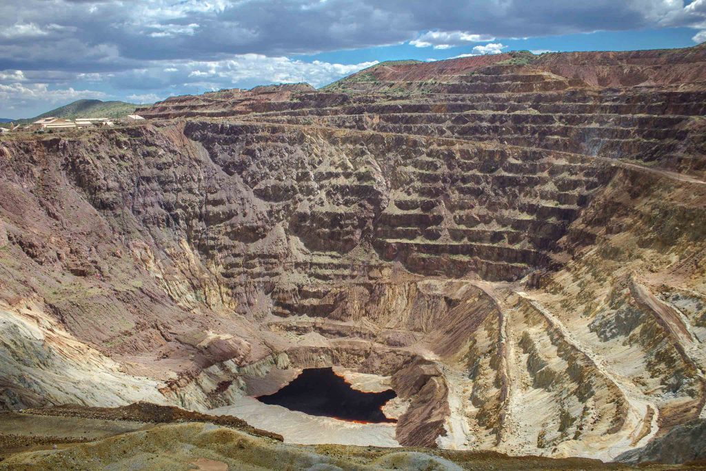 A terraced rock pit with dark red liquid in the bottom and blue sky with clouds above.