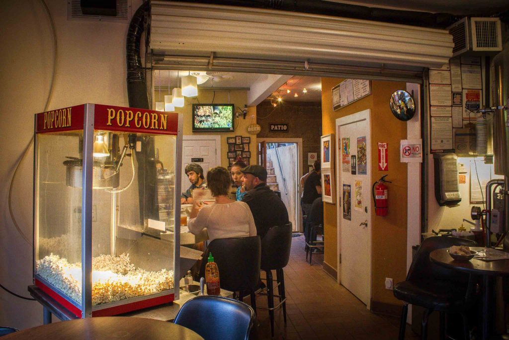 People sitting along a bar, behind high-top tables and a popcorn machine.