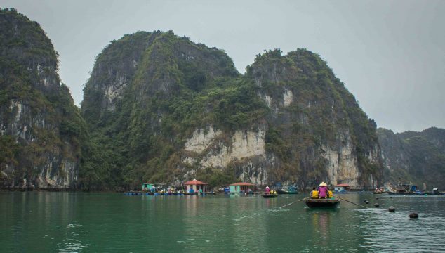 Boats rowing toward small overwater buildings, in front of tree-covered karst mountains and a gray sky.