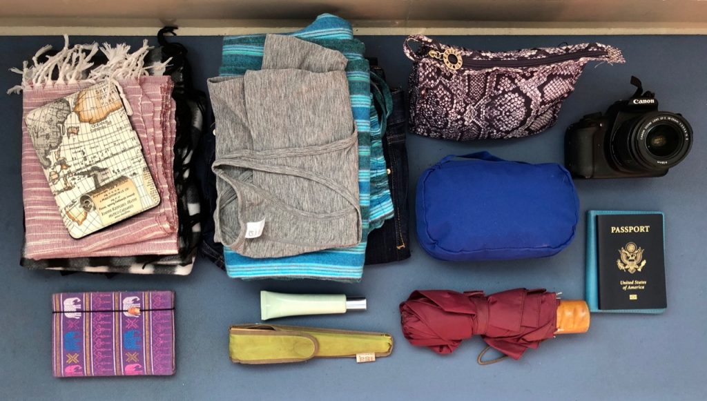 Rows of small bags, piles of folded clothes, and items like a notebook, camera, umbrella, and U.S. passport, on a blue background.