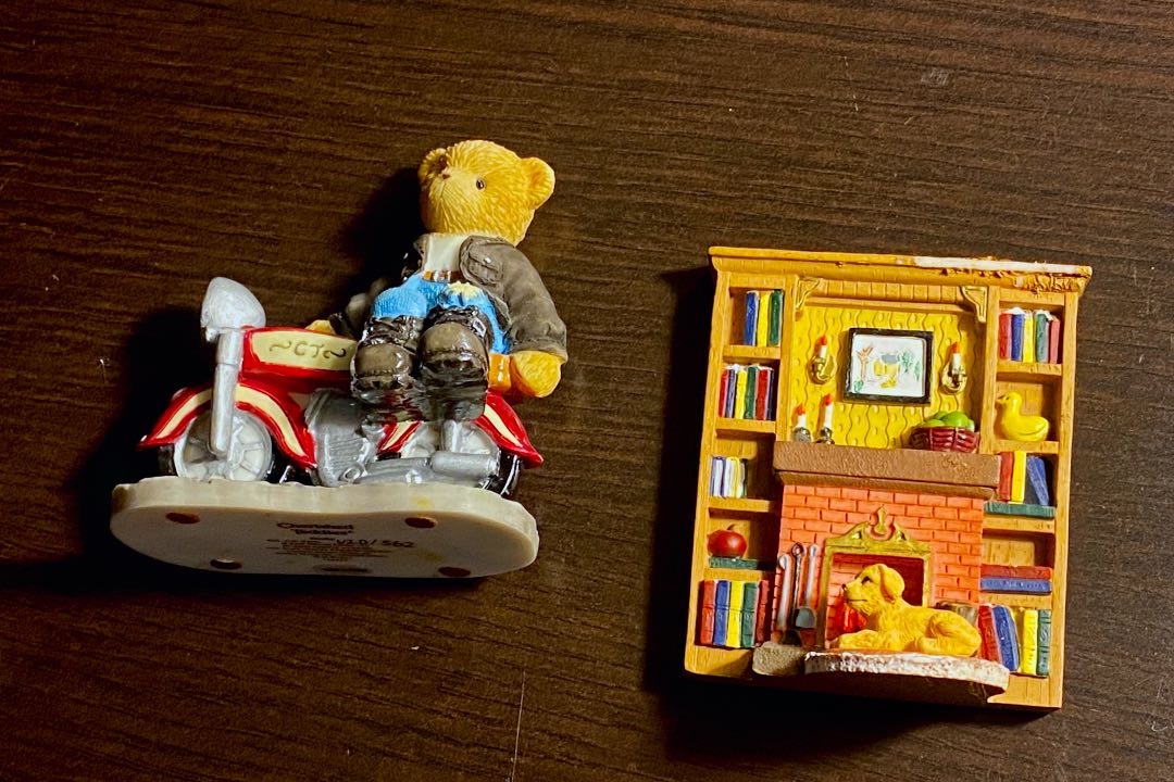 Two christmas ornaments on a wooden table, a teddy bear on a motorcycle and a dog sitting in front of a fireplace and bookshelf.