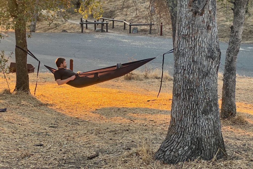 Man laying in a black hammock hung between two trees, looking toward a road and dirt path.
