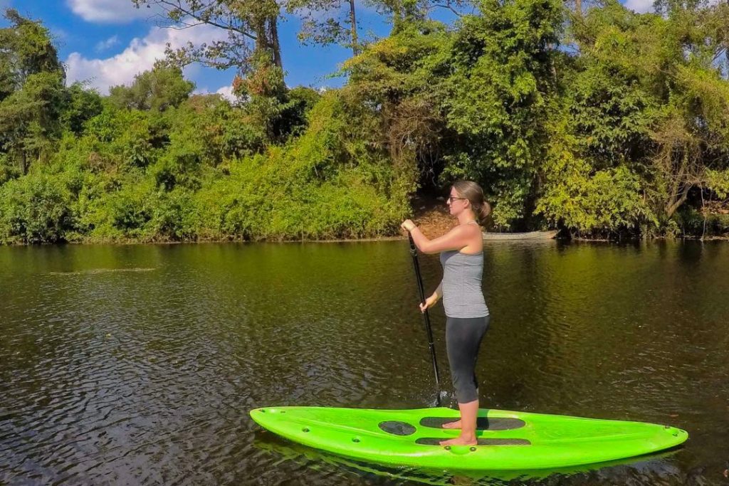 Woman paddling a neon green stand-up paddleboard in front of a tree-lined riverbank.
