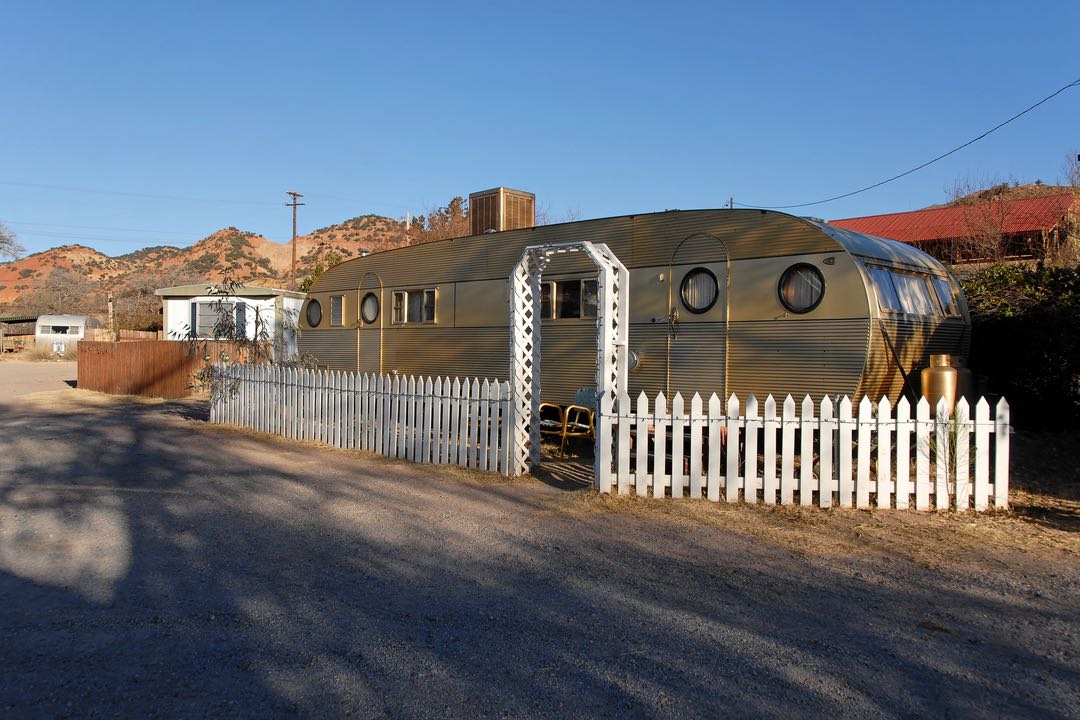 A modern, Airstream-style trailer sits behind a white picket fence with low hills in the background.