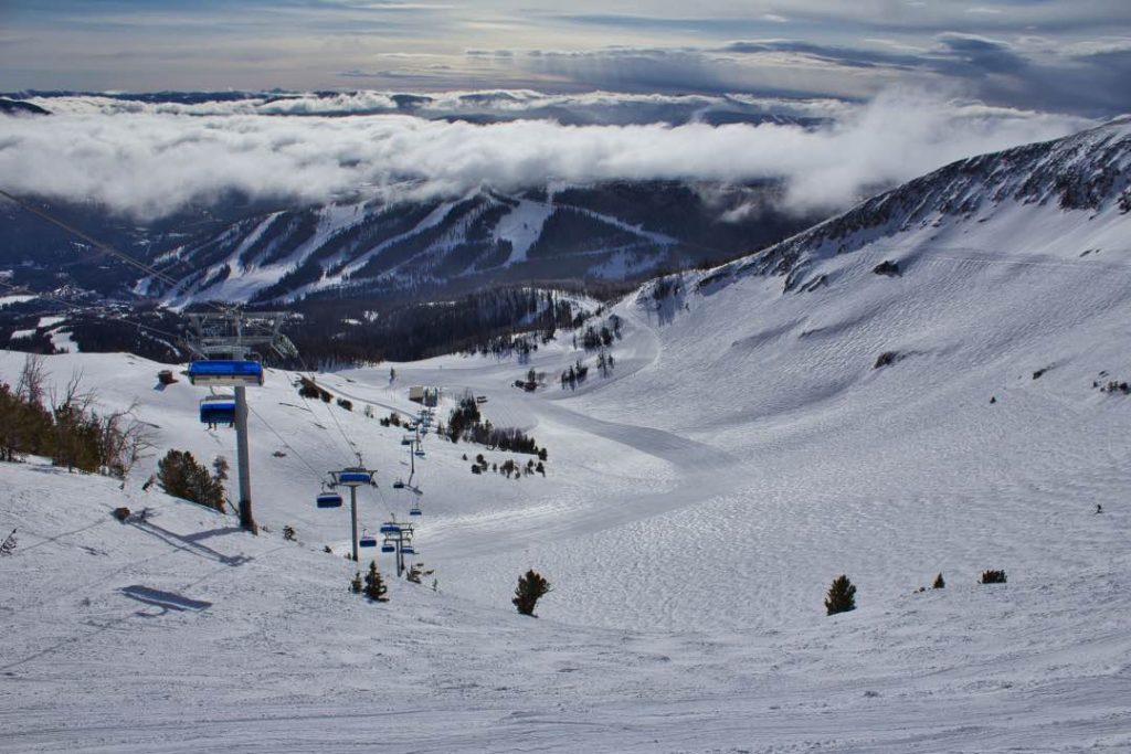 Looking down a ski run, with a chairlift running along the left side and cloud-shrouded mountains in the background.