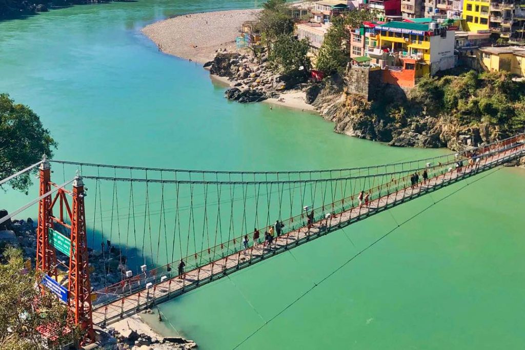 People walking on a pedestrian bridge over a bright green river, with colorful buildings and a beach on the right bank.