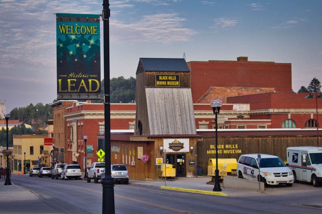 Downtown street with a banner reading "Welcome Historic Lead" on one side and a wooden building with signs reading "Black Hills Mining Museum" on the other.