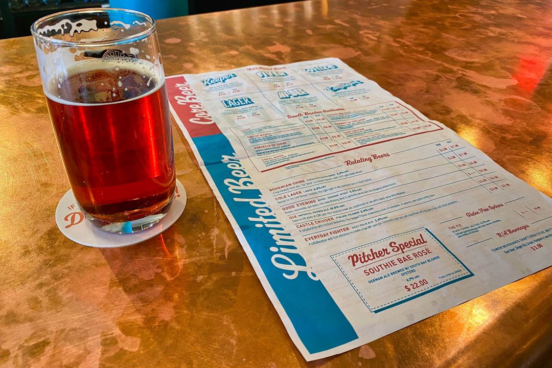 A pint glass filled with red-hued beer sits on a copper-plated bar next to a red and blue paper beer menu.