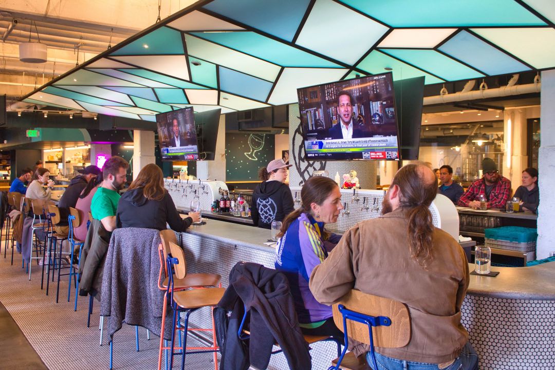 Several people sit alongside a bar with modern aesthetics while a flat screen TV above them plays sports news.