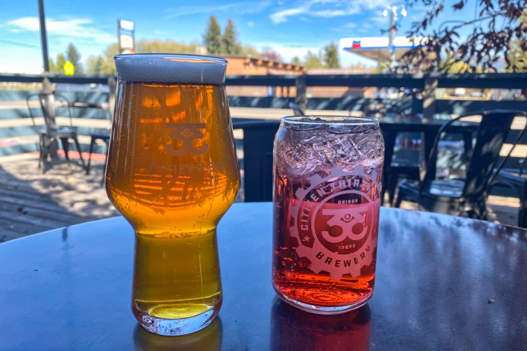A tulip shaped glass filled with amber-hued beer sits next to a straight-walled glass of light red liquid both bearing a Citizen 33 gear-shaped logo and sitting on a metal table outside.