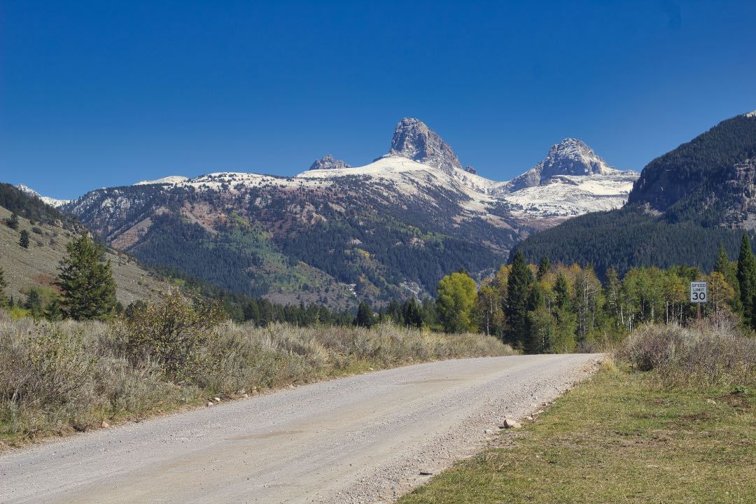 A set of mountains with two very prominent, rocky peaks, sit in front of a forested gravel road.