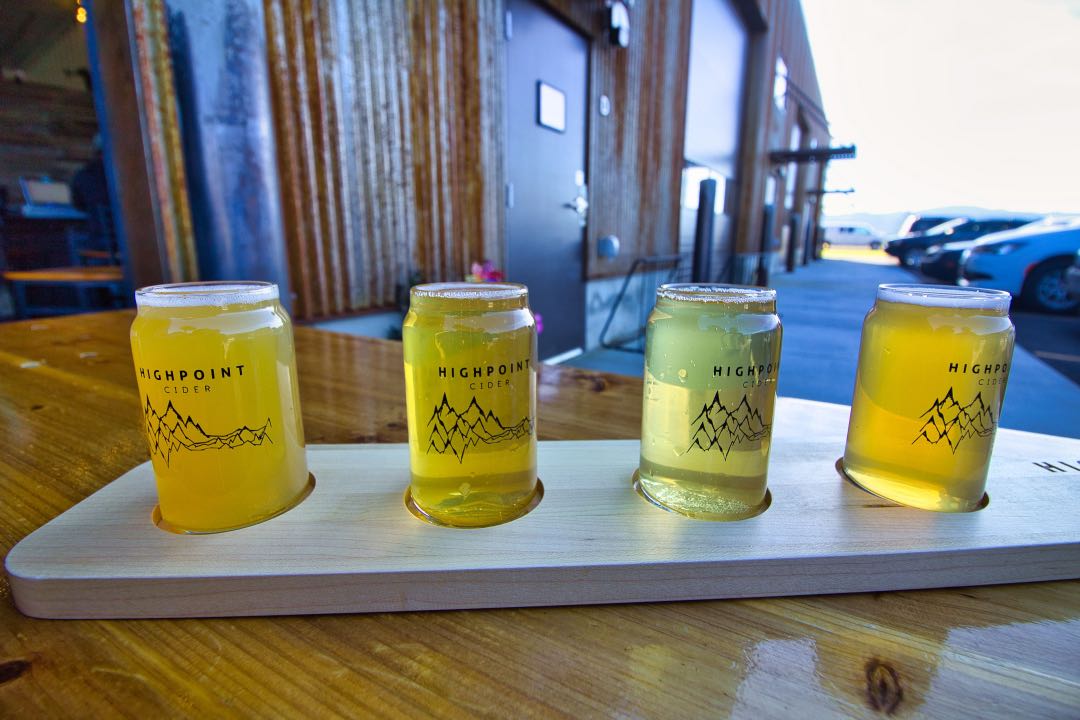 Four small glasses of pale yellow liquid sit in round cutouts on a board laid across a dining table.