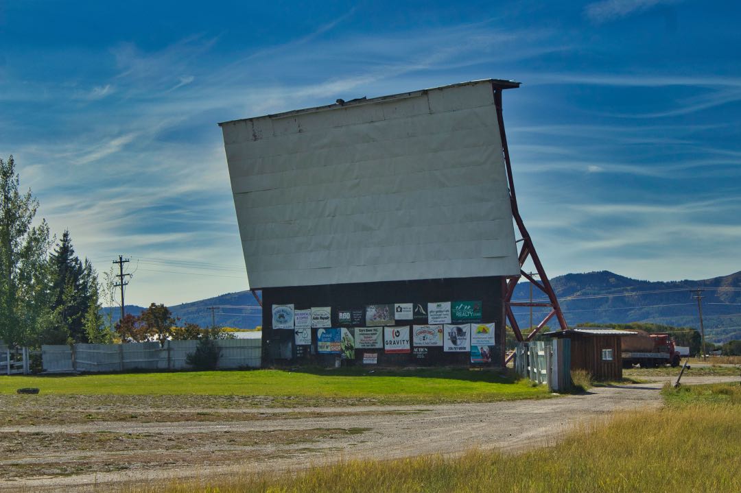 A large white fabric movie screen sits behind a empty grassy parking lot with low mountains in the background.
