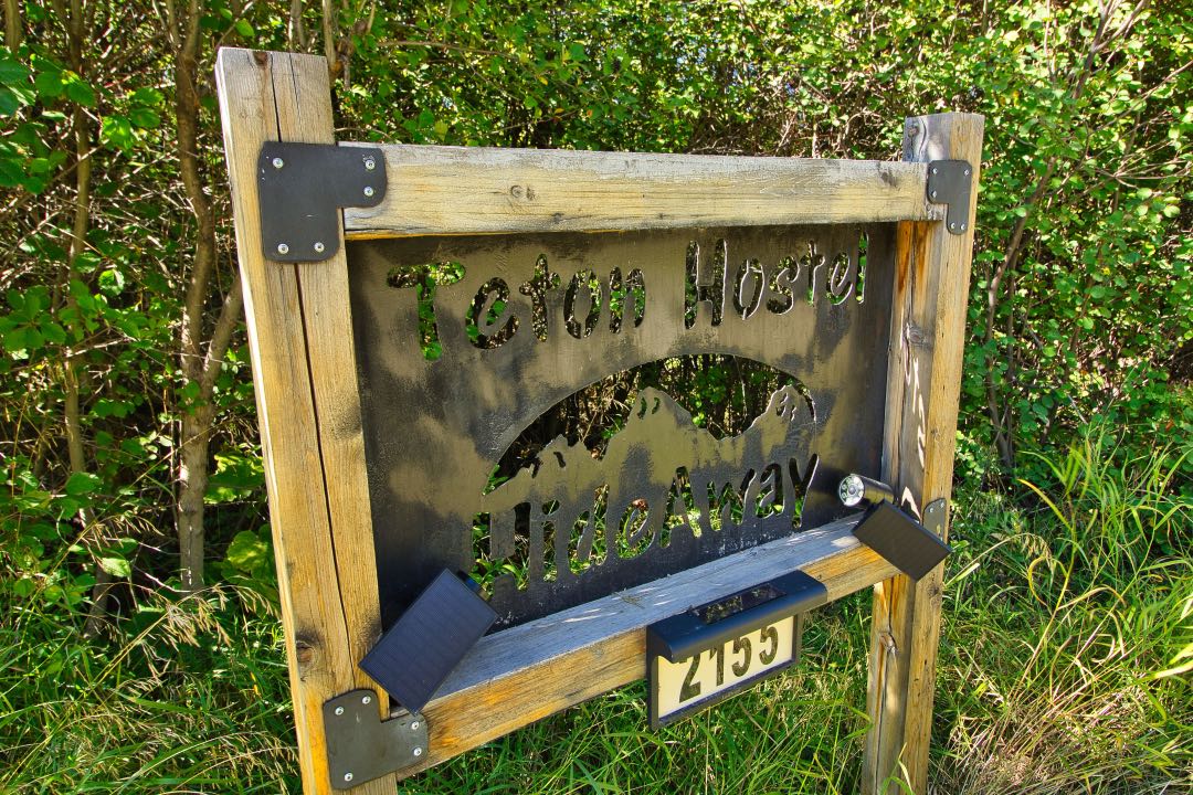 Rustic metal sign with wooden frame stating Teton Hideaway Hostel against a backdrop of green shrubs.