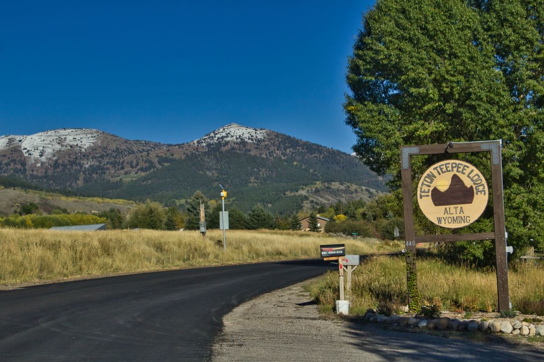 Asphalt road leading past a circular wooden sign saying Teton Teepee Lodge with a single snow-capped mountain int he background.