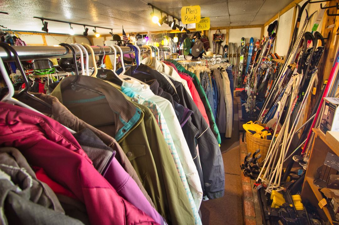 A rack of multicolored winter coats with sets of old skis and camping equipment in the background.