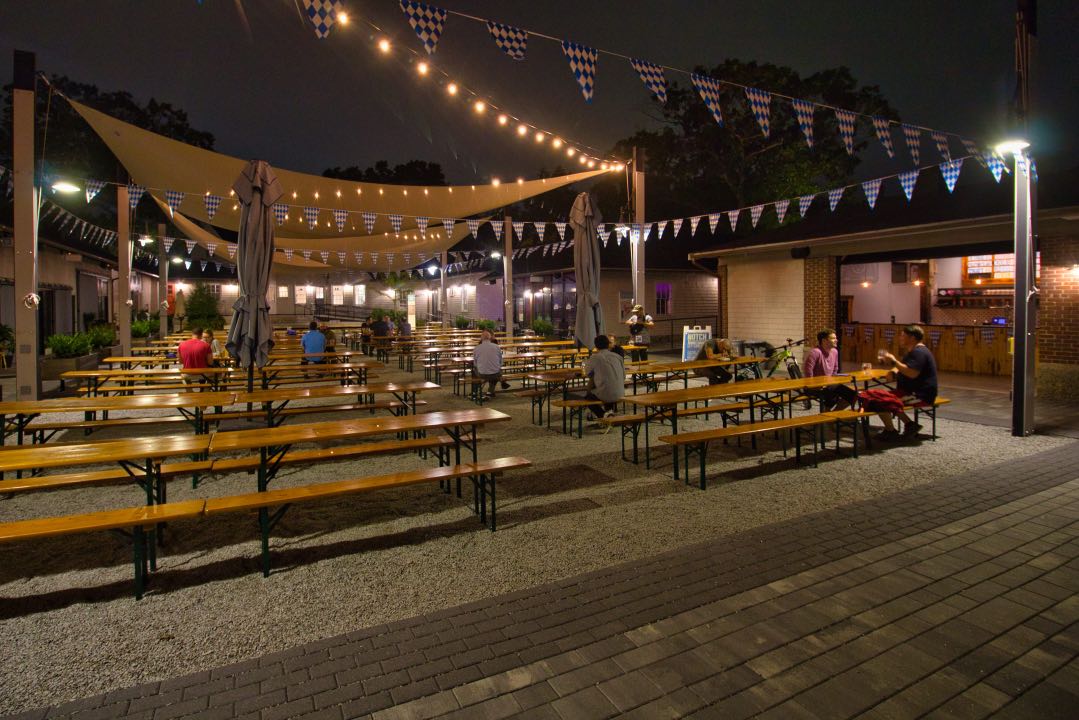Several rows of long wooden tables sit in a courtyard with a few customers sitting at them. A string of triangular flags and lights hang above them under a dark night sky.