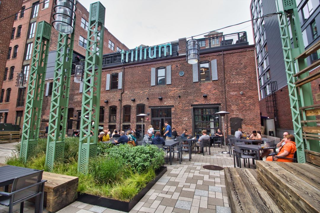 Dozens of customers sit at dark wooden tables on an outdoor patio with a rustic brick building behind them and a large light blue sign saying "Trillium" hanging above them