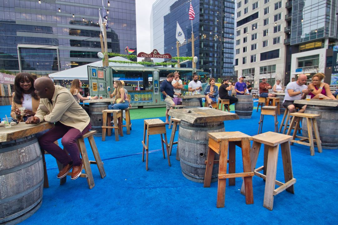 Couples and friends sit on rustic wood stools at tables made from barrels. In the background is a wall of metal and glass skyscrapers overlooking the beer garden space.
